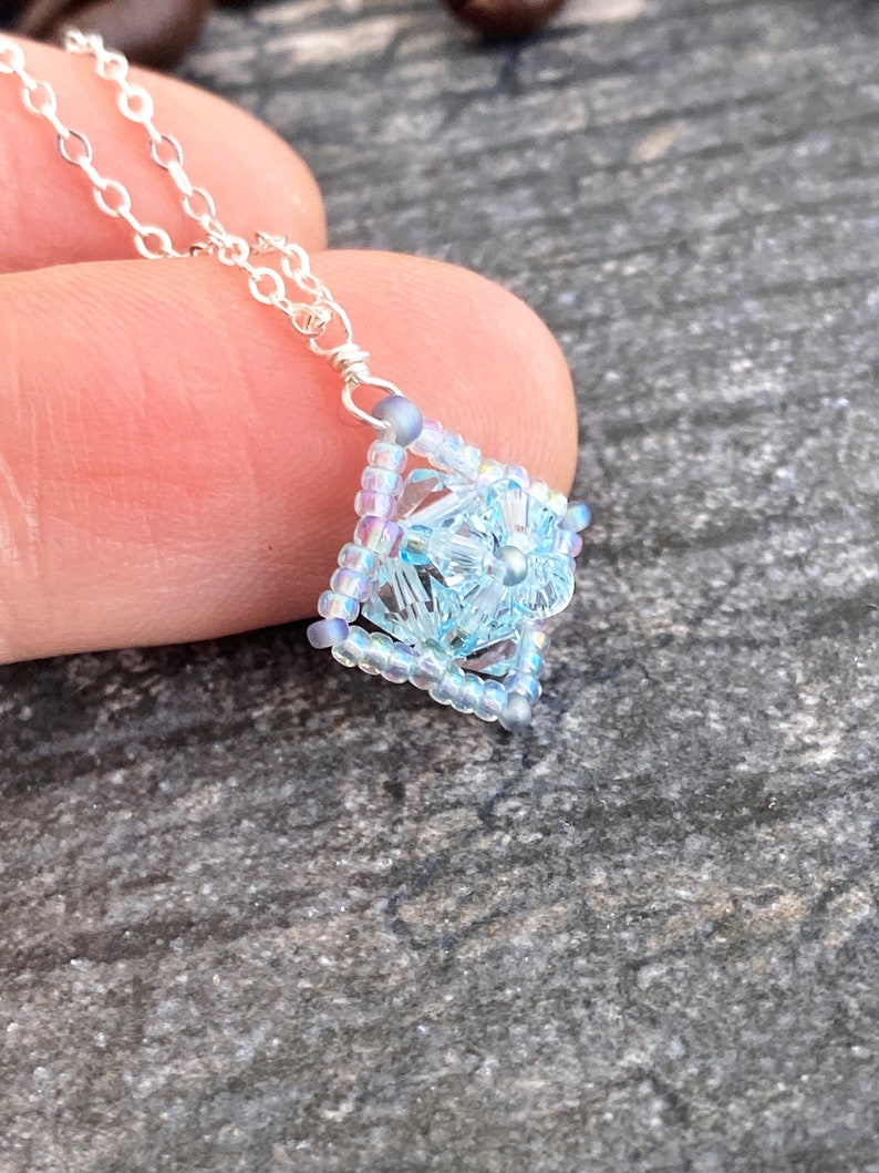 May include: A delicate silver chain necklace with a light blue and iridescent beaded pendant in the shape of a square.