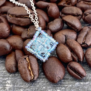 May include: A delicate silver chain necklace with a light blue beaded pendant shaped like a square. The pendant has a floral design in the center and is surrounded by iridescent beads.