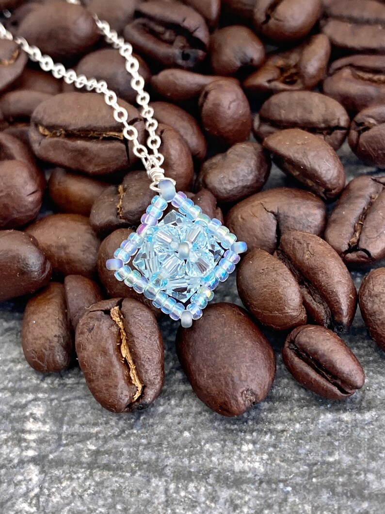 May include: A delicate silver chain necklace with a light blue beaded pendant shaped like a square. The pendant has a floral design in the center and is surrounded by iridescent beads.