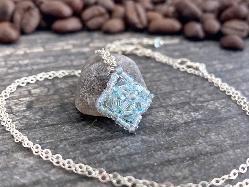May include: A delicate silver chain necklace with a light blue crystal pendant. The pendant is a square shape and is made of small, faceted crystals.