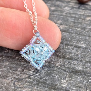 May include: A delicate silver chain necklace with a light blue iridescent square pendant made of beads.