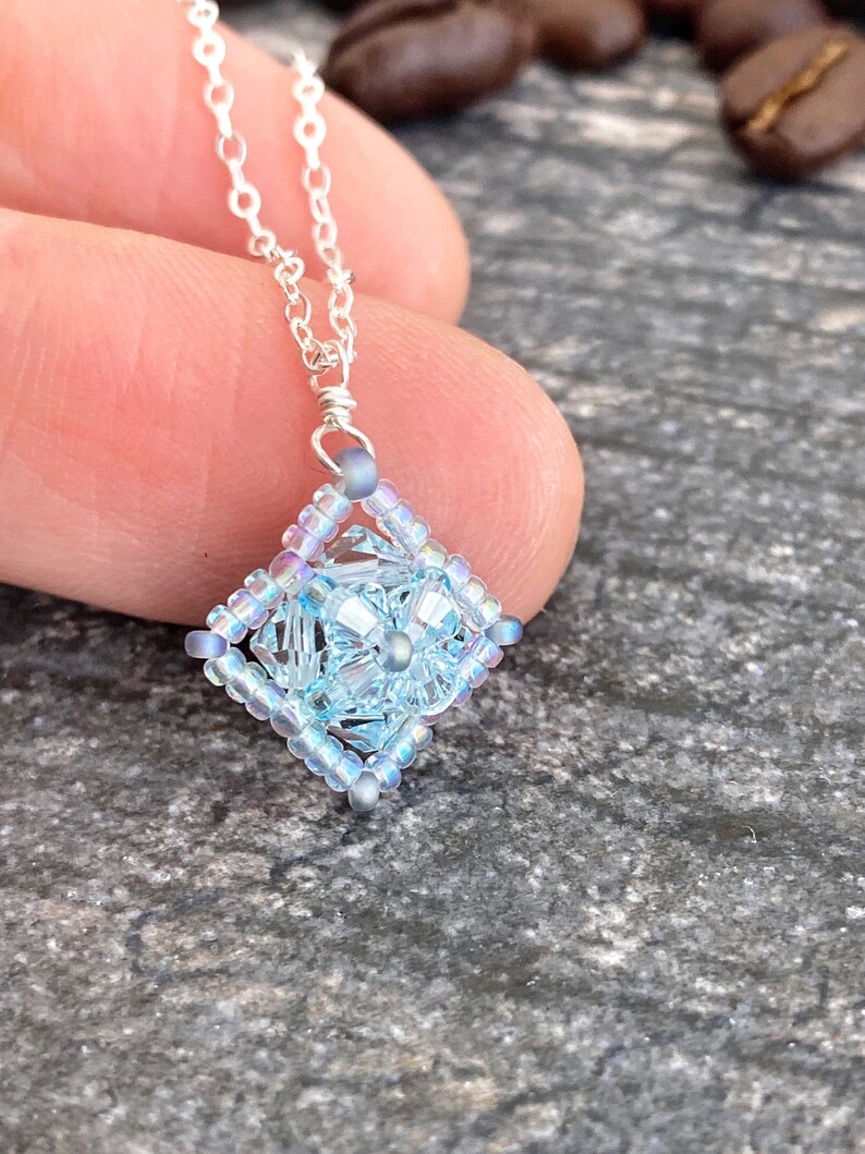 May include: A delicate silver chain necklace with a light blue iridescent square pendant made of beads.