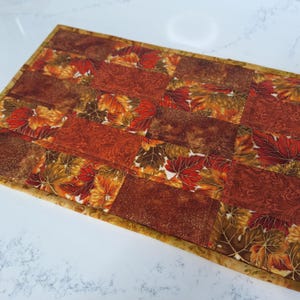 May include: A rectangular table runner with a patchwork design. The runner features a repeating pattern of rectangular blocks with autumn leaf and swirl patterns in shades of orange, red, and brown. The edges are trimmed with a gold border.