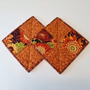 May include: Two square quilted fabric coasters. Each coaster features a sunflower design with red, yellow, and green petals on a black background. The borders and backing are a solid orange with a leaf pattern.