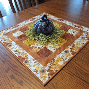 May include: A square fabric table topper featuring a patchwork center of brown and gold squares, surrounded by a border of autumn leaves and flowers in shades of orange, yellow, and white. A dark purple decorative pumpkin sits on green foliage.