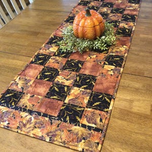 May include: A quilted table runner with a black and brown checkered pattern featuring autumn leaves and vines. The runner has a border of orange, brown, and gold leaves. A small ceramic pumpkin sits on top of the runner.