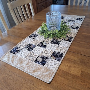 May include: A quilted table runner with a patchwork design featuring floral and paisley patterns in beige, navy, and cream. A clear glass candle holder sits atop a green wreath in the center of the runner.