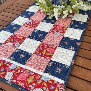 May include: A rectangular quilted table runner featuring a patchwork design of red, white, and navy blue fabric squares. The pattern includes floral prints, small white cherries, and solid-colored squares. A ceramic candle holder sits on top.