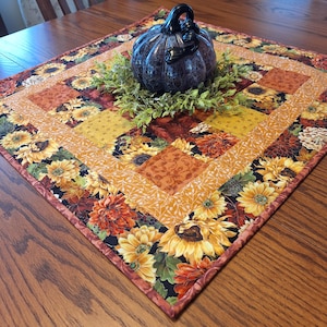 May include: A square fabric table topper featuring a patchwork design of sunflowers, autumn leaves, and floral patterns in shades of orange, yellow, and brown. A dark, glossy, textured decorative pumpkin sits in the center on a green leaf wreath.