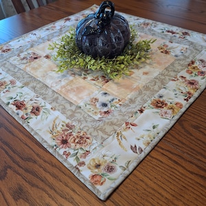 May include: A square fabric table topper featuring a floral print border with shades of orange, brown, and cream. The center has a beige patterned section. A dark purple glass pumpkin sits atop a green artificial wreath in the middle.