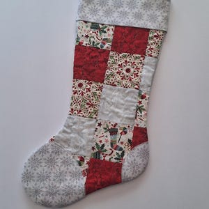 May include: A quilted Christmas stocking with a patchwork design. The stocking features red, white, and silver fabrics with snowflake, floral, and holiday-themed patterns. A loop is attached for hanging.