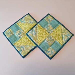 May include: Two square quilted potholders. Each potholder is made of light green and teal fabric with floral patterns. The potholders are approximately 8 inches square and have a dark blue border.