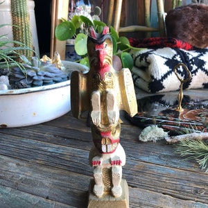 Könnte beinhalten: Eine Holzfigur eines Totempfalses mit goldener und roter Bemalung. Der Totempfahl zeigt einen Vogel mit Schnabel und eine menschenähnliche Figur mit einem Gesicht.