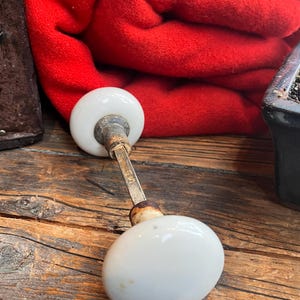 Peut inclure: Une paire de poignées de porte vintage en porcelaine blanche avec une tige en métal terni. Les boutons ovales reposent sur une surface en bois vieilli, avec une couverture rouge pliée en arrière-plan.