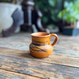 May include: A small, handmade, orange-brown ceramic mug with a handle. The mug has dark brown, hand-painted stripes and a rounded body. The mug is on a weathered, wooden surface. The mug is likely used for drinking beverages.