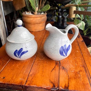 May include: A ceramic sugar bowl with a lid and a creamer jug. Both are speckled grey with blue floral designs. The sugar bowl has a small knob on the lid. The creamer has a handle and a spout. The set is on a wooden surface.