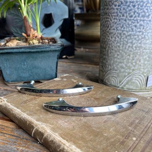 Peut inclure: Deux poignées en métal argenté brillant, de forme incurvée, avec de petits boutons arrondis. Elles reposent sur un livre ancien à la couverture beige usée. L'image évoque des éléments de décoration intérieure anciens ou vintage.
