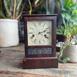 Peut inclure: Une horloge ancienne en bois avec un cadran carré et des chiffres romains. L'horloge a un cadre marron foncé et un panneau décoratif bleu et or en bas. L'horloge est posée sur une surface en bois.