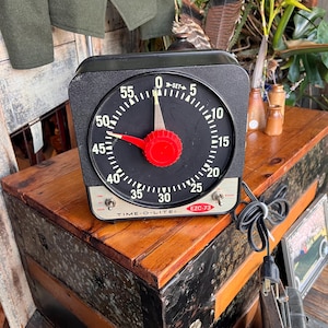 以下が含まれることがあります： 黒い文字盤、白い数字、赤いダイヤルが付いた、黒いTime-O-Lite EZC-73タイマー。タイマーは木製の表面にあります。タイマーの文字盤には「TIME-O-LITE」と「EZC-73」の文字があります。