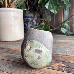 May include: A small, rounded ceramic vase with a dark grey glaze and a green and white glaze on one side. The vase is sitting on a wooden surface.