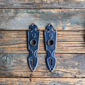 May include: A pair of dark blue antique door handle backplates with keyhole and circular knob openings. The ornate backplates are set against a weathered wooden background, showing signs of age and use.