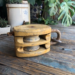 Peut inclure: Poulie en bois vintage avec une finition de peinture jaune vieillie. Le bloc comporte deux roues rainurées et une grande boucle en métal. L'article est probablement utilisé pour soulever des objets lourds. La poulie est posée sur une surface en bois rustique.