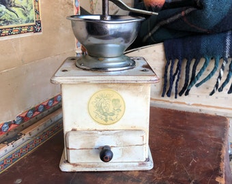 May include: An antique coffee grinder with a white painted wooden base and a silver metal top. The grinder features a drawer with a black knob and a decorative emblem. The wooden handle is brown. The grinder sits on a brown surface.