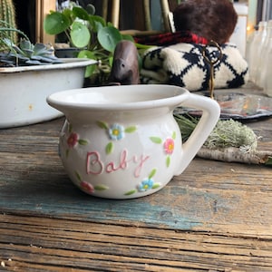 May include: White ceramic mug with a floral design and the word "Baby" in pink lettering. The mug has a handle and is sitting on a wooden surface.