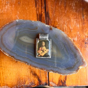 May include: A rectangular silver pendant with a small image of a woman holding a baby. The pendant is set on a blue and gray agate slice, resting on a wooden surface.