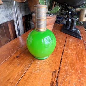 Op de afbeelding: Een heldergroene glazen lamp met een ronde basis en een cilindrische metalen bovenkant. De lamp heeft een glanzende afwerking en staat op een houten oppervlak. De metalen bovenkant heeft een messing accent.