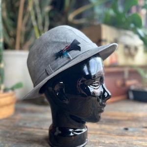 May include: A grey fedora hat with a small brim and a decorative feather accent. The hat is displayed on a black mannequin head. The hat has a band around the crown and a small feather accent.