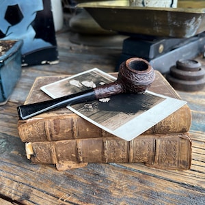 Puede incluir: Una pipa de tabaco vintage con un cuenco texturizado y una boquilla negra descansa sobre una pila de libros antiguos. Dos fotografías en blanco y negro están colocadas encima de los libros. El fondo incluye una balanza y una pequeña planta en maceta.