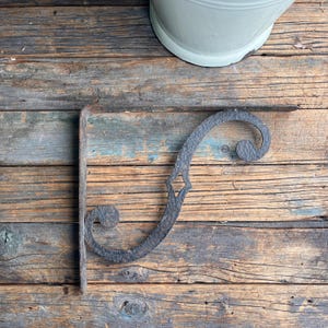 Può includere: Una staffa per mensola in ferro battuto grigio scuro con un design a volute decorativo. La staffa presenta un ritaglio a forma di diamante. È montata su una superficie di legno invecchiato.