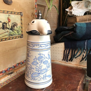 Peut inclure: Une chope en céramique blanche avec un motif floral bleu et blanc. La chope a un couvercle avec un visage sculpté et un bec.