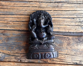 Op de afbeelding: Een donkerbruin, ingewikkeld gesneden beeld van de hindoe-godheid Ganesha. De sculptuur toont Ganesha zittend, met meerdere armen, een olifantenkop en een grote buik. Het beeld staat op een decoratieve basis.