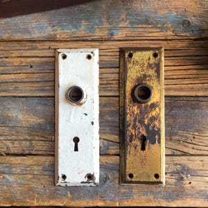 Peut inclure: Deux plaques de porte anciennes, l'une blanche et l'autre dorée, avec un trou de serrure et un trou rond pour un bouton. Les deux plaques ont un aspect usé et vieilli.