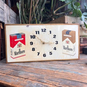May include: Rectangular wall clock with a cream-colored face, featuring black numerals and gold-colored hands. The clock face has images of Marlboro cigarette packs on either side. The clock is framed in a light brown wooden border.