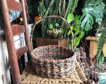 May include: A woven wicker basket with a brown handle sits on a wooden chair with a woven straw seat. The basket is filled with colorful yarn. The chair is in front of a wall of green plants.