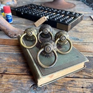 Puede incluir: Cuatro tiradores de cajón antiguos de latón con asas de anillo descansan sobre un libro antiguo. Los tiradores tienen un diseño floral decorativo alrededor del pomo central. El libro es verde con letras doradas. Un ábaco está en el fondo.