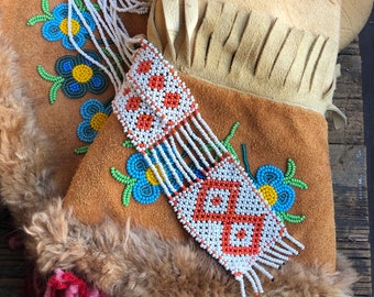 以下が含まれることがあります： 白とオレンジのビーズネックレスが、青と緑の花柄の刺繍が施された茶色の革製のポーチからぶら下がっている。