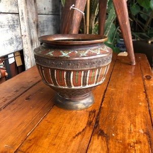 May include: An antique bronze-coloured vase with a wide rim and a decorative band featuring a repeating circular pattern. The body of the vase is adorned with vertical stripes in red, green, and white. The vase sits on a wooden surface.