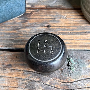May include: A black gear stick knob with white markings for gears: L, 1, 2, 3, N, and R. The spherical knob is made of a smooth, shiny material. It rests on a rustic, weathered wooden surface.