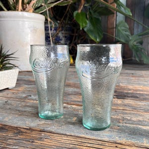 Könnte beinhalten: Zwei transparente Coca-Cola-Gläser mit strukturierter Oberfläche. Die Gläser haben eine hellgrüne Tönung am Boden und tragen das Coca-Cola-Logo. Die Gläser stehen auf einer verwitterten Holzoberfläche.