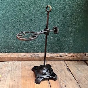 May include: An antique, cast iron laboratory stand with a circular holder and a ring at the top. The stand has a black, ornate base and a dark, aged finish. The holder is designed to cradle a round-bottom flask or other glassware.