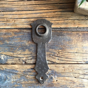 May include: A black metal door handle with a decorative design. The handle has a round opening in the centre and a pointed bottom.