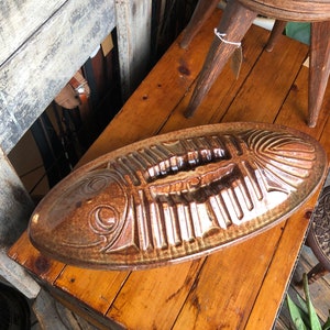 May include: A brown ceramic fish-shaped serving dish with a detailed fish skeleton design. The dish is oval and rests on a wooden surface. The dish is a decorative item.