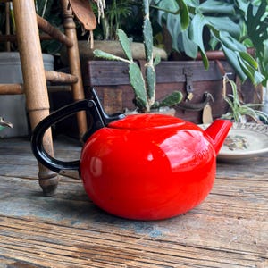 May include: A vibrant red teapot with a black handle and lid. The teapot has a rounded shape and a spout. The handle is attached to the side of the teapot. The teapot is sitting on a wooden surface.