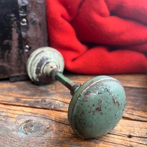 May include: A vintage green and white door knob with a distressed finish rests on a weathered wooden surface. A red blanket is in the background, along with a dark metal object. The knob has a round shape.