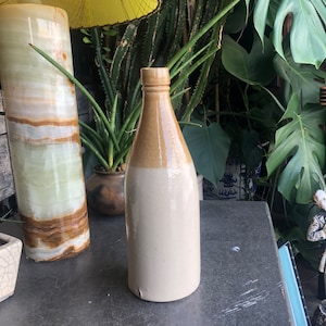 Peut inclure: Une bouteille en grès vintage avec un design bicolore. La partie supérieure est brun clair, passant à une base de couleur crème. La bouteille a un col étroit et un corps arrondi, probablement utilisée pour stocker des liquides. La bouteille mesure environ 25 cm de haut.