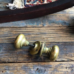 Może przedstawiać: Dwa antyczne mosiężne gałki drzwiowe o okrągłym kształcie i ozdobnym wzorze. Gałki leżą na drewnianej powierzchni.